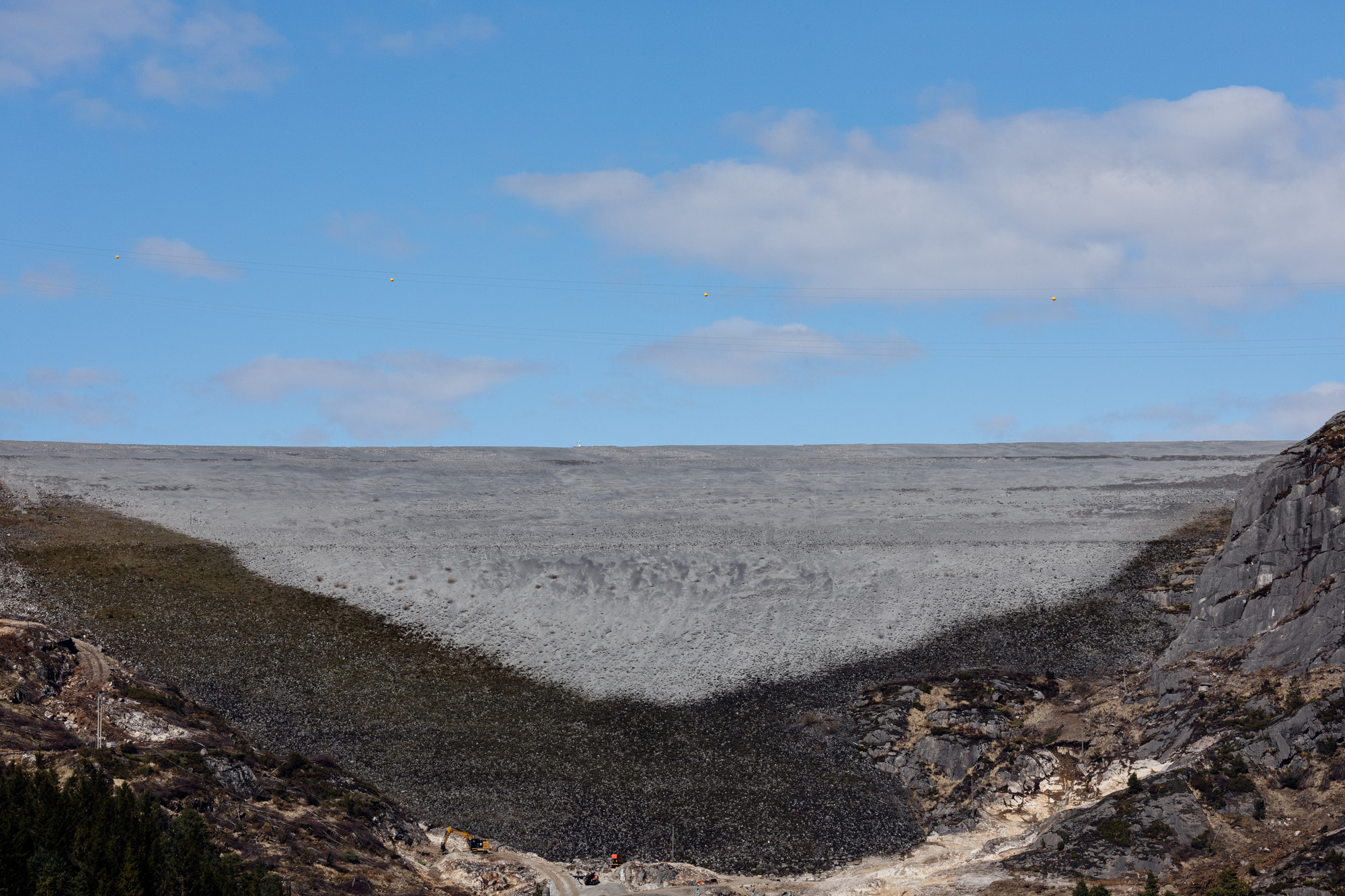 A landscape image of a large grey mine and a bright blue sky with a few clouds.