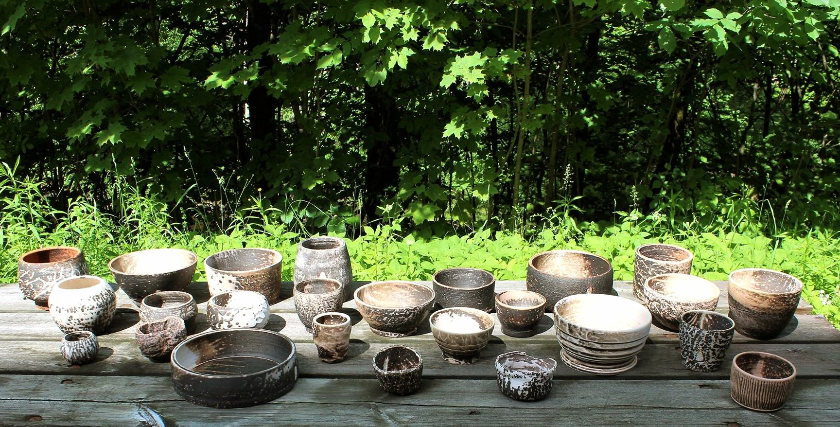 An outside table with 25 different sized and shaped brown and white ceramic bowls. In the background bright green plants and trees.