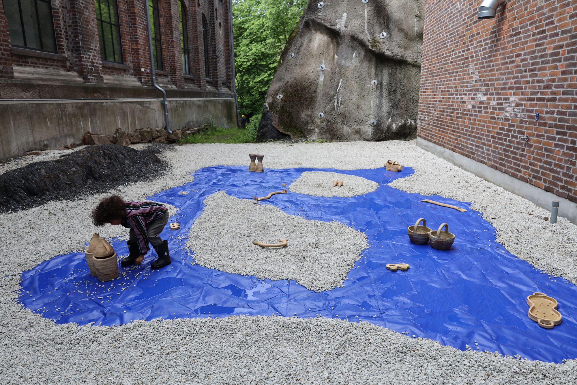 En installation som består av keramikkobjekter på et delvis grusbelagt blått underlag. I forgrunnen er det et litet barn som ser på en av skulpturene.