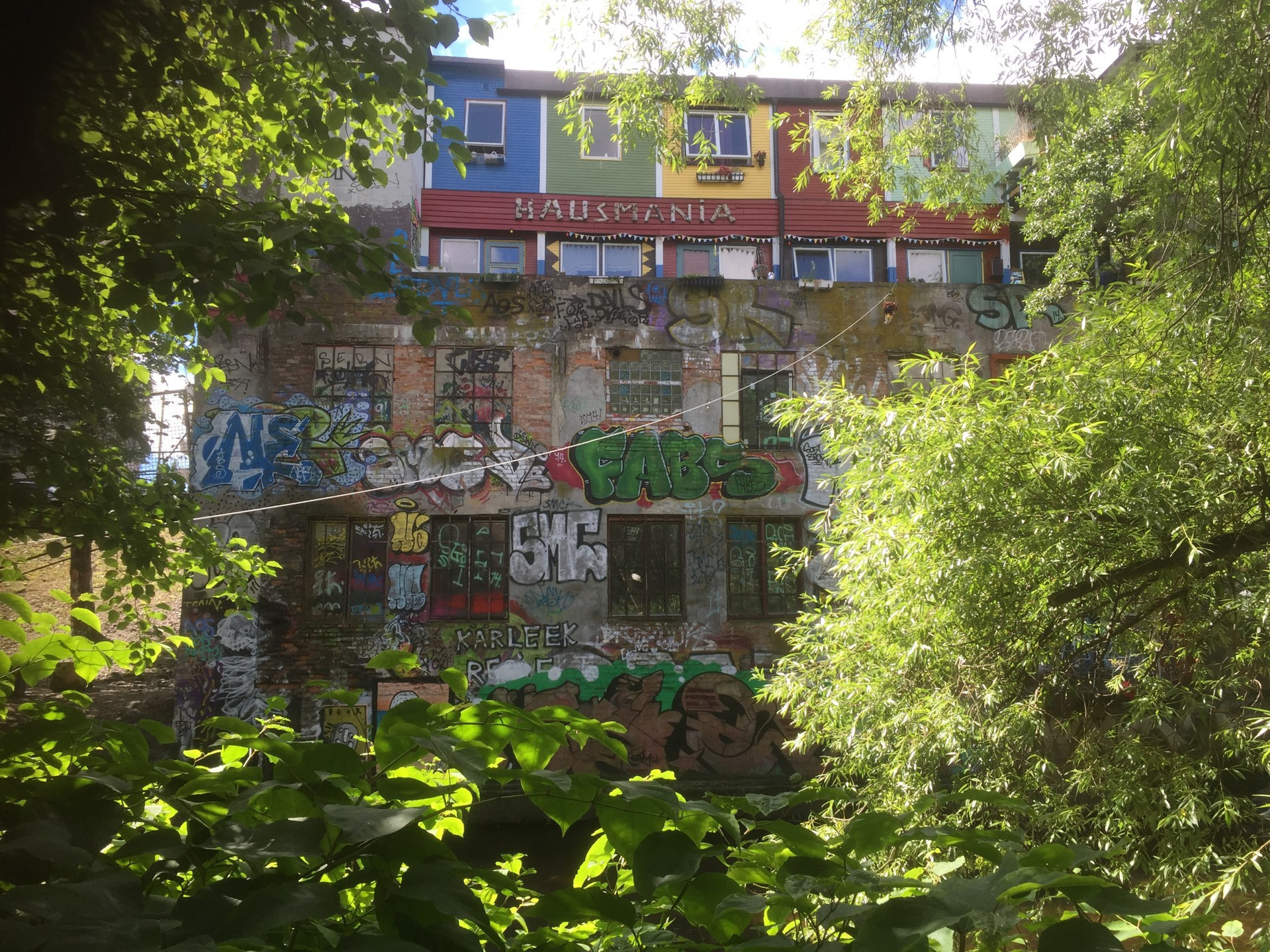 Colour photo of the backside of Hausmania culture house with a wall covered in graffiti. Seen through green tree branches.
