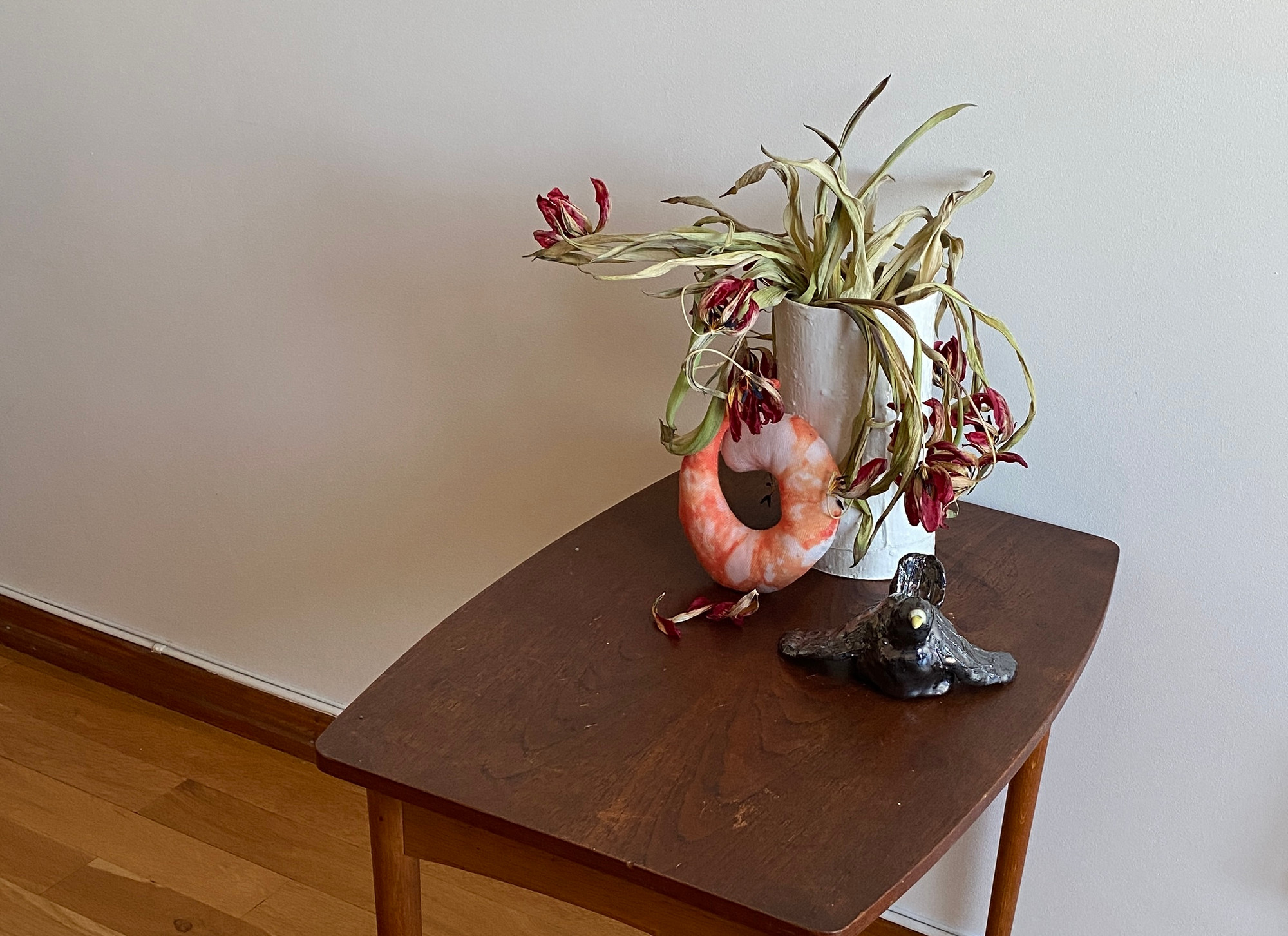 Photo of a small table with a vase of dried tulips, a soft toy shaped like a shrimp and a black ceramic bird.