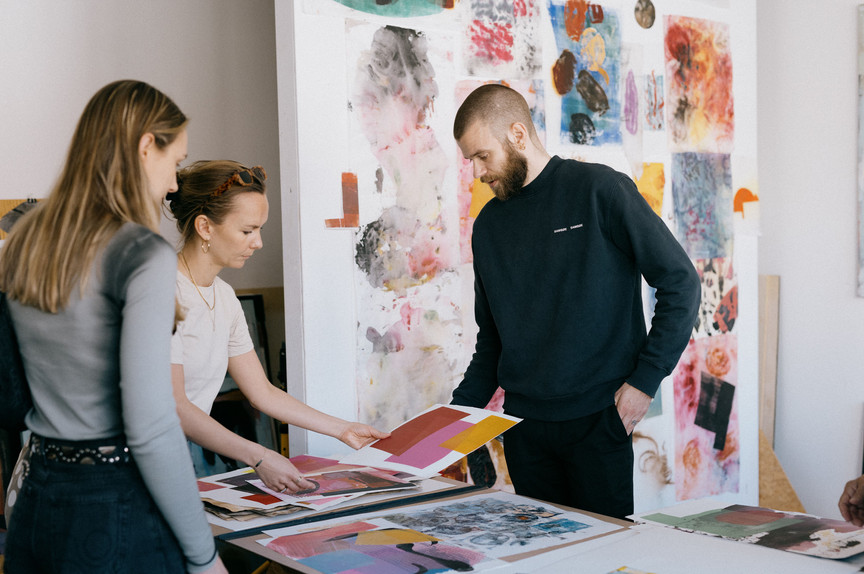 Three people looking at colourful art works on a table.