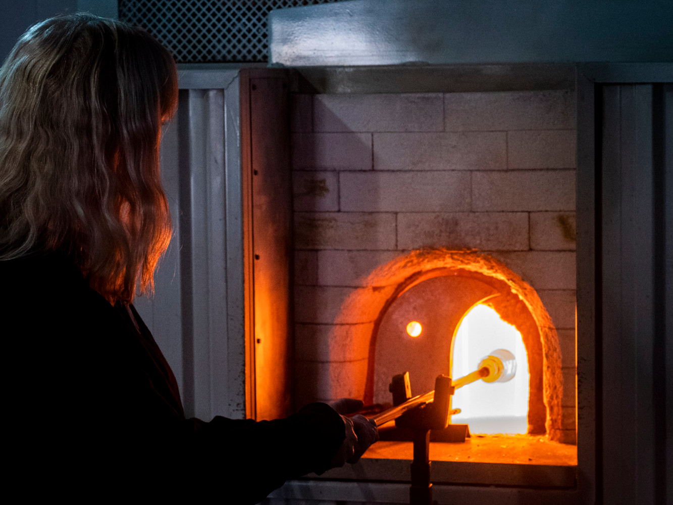 A colour photo of a long haired person who warms a stick with glass at the end in a glowing hot oven.