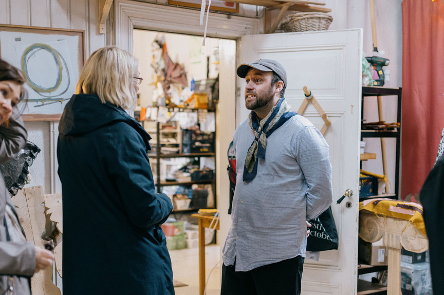 Artist Fredrik Floen, wearing a cap and silk scarf, greets two visitors in his studio.