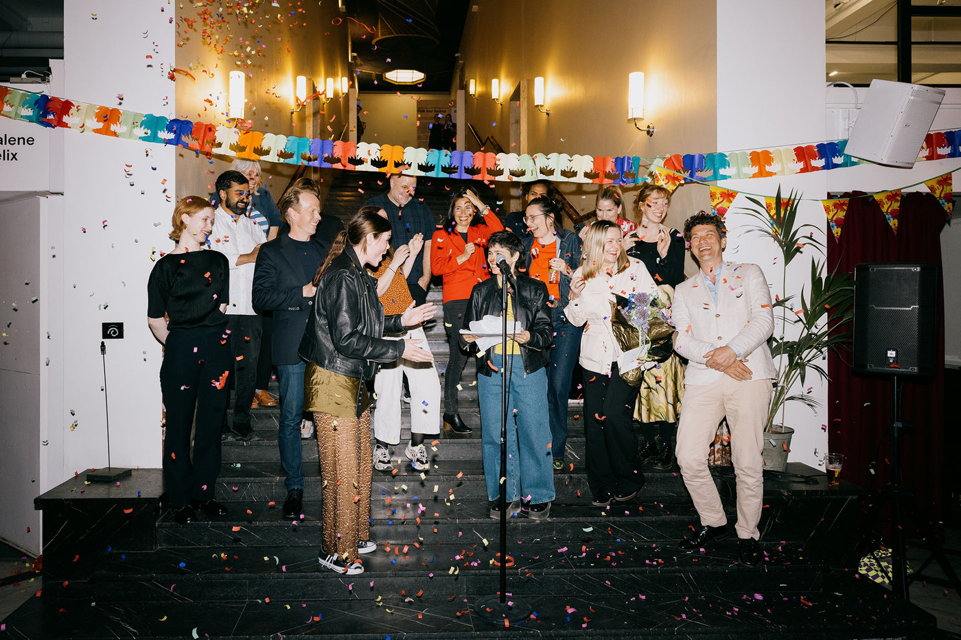 Fifteen representatives from Atelier Kunstnerforbundet is pictured in a staircase behind a microphone, in a cloud of confetti.