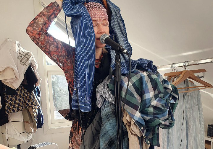 Turud Alida singing into a microphone with closed eyes. With her left hand she is holding a pile of clothes and with her right hand she is holding a blue striped shirt and letting it hang above her head.
In the background, from left to right, there is a mannequin with several layers of clothes, a window and a clothes rack with a shirt and empty hangers hanging from it.