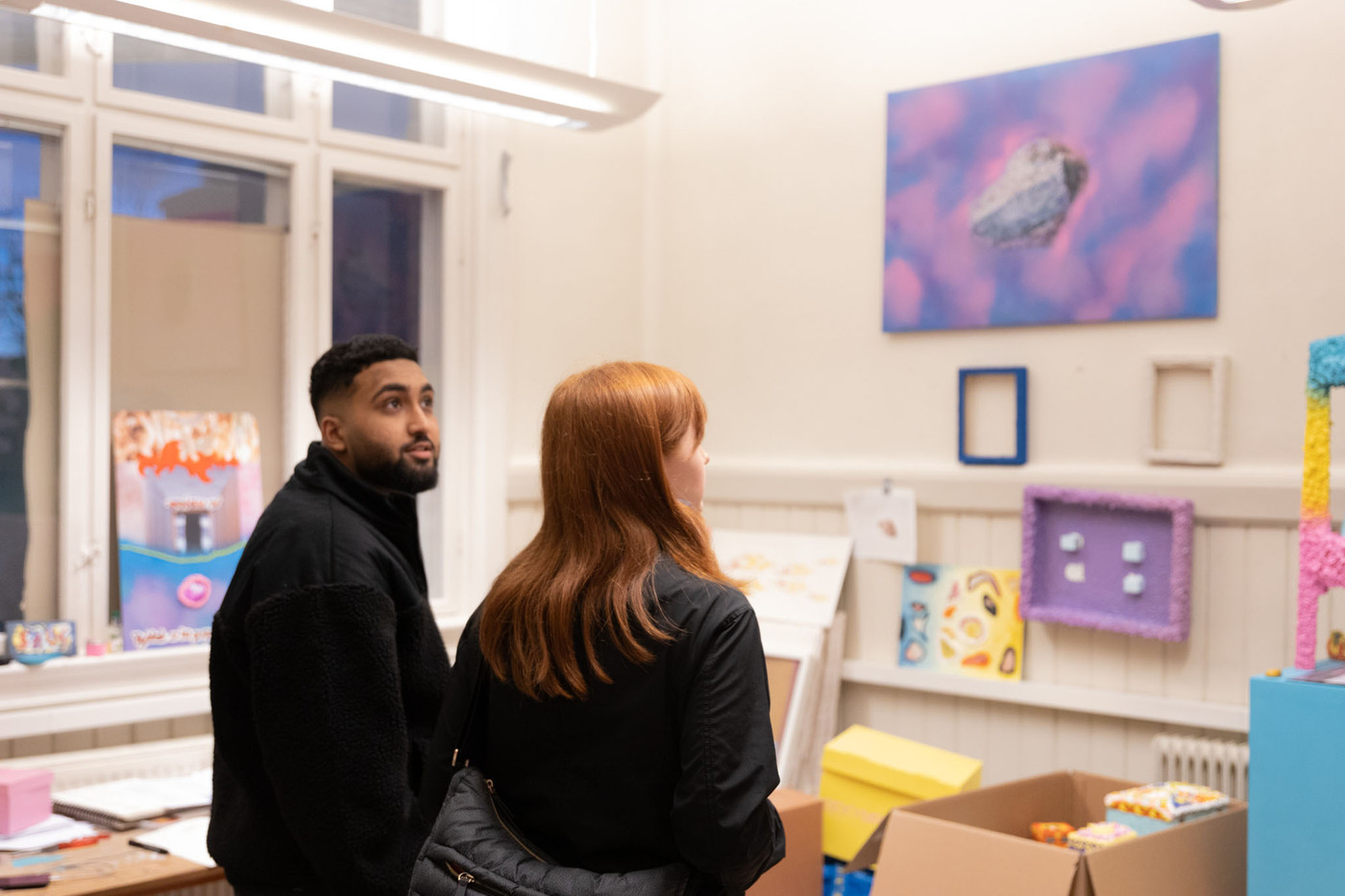 Two people look around a room filled with artworks in bright yellow, pink and purple.
