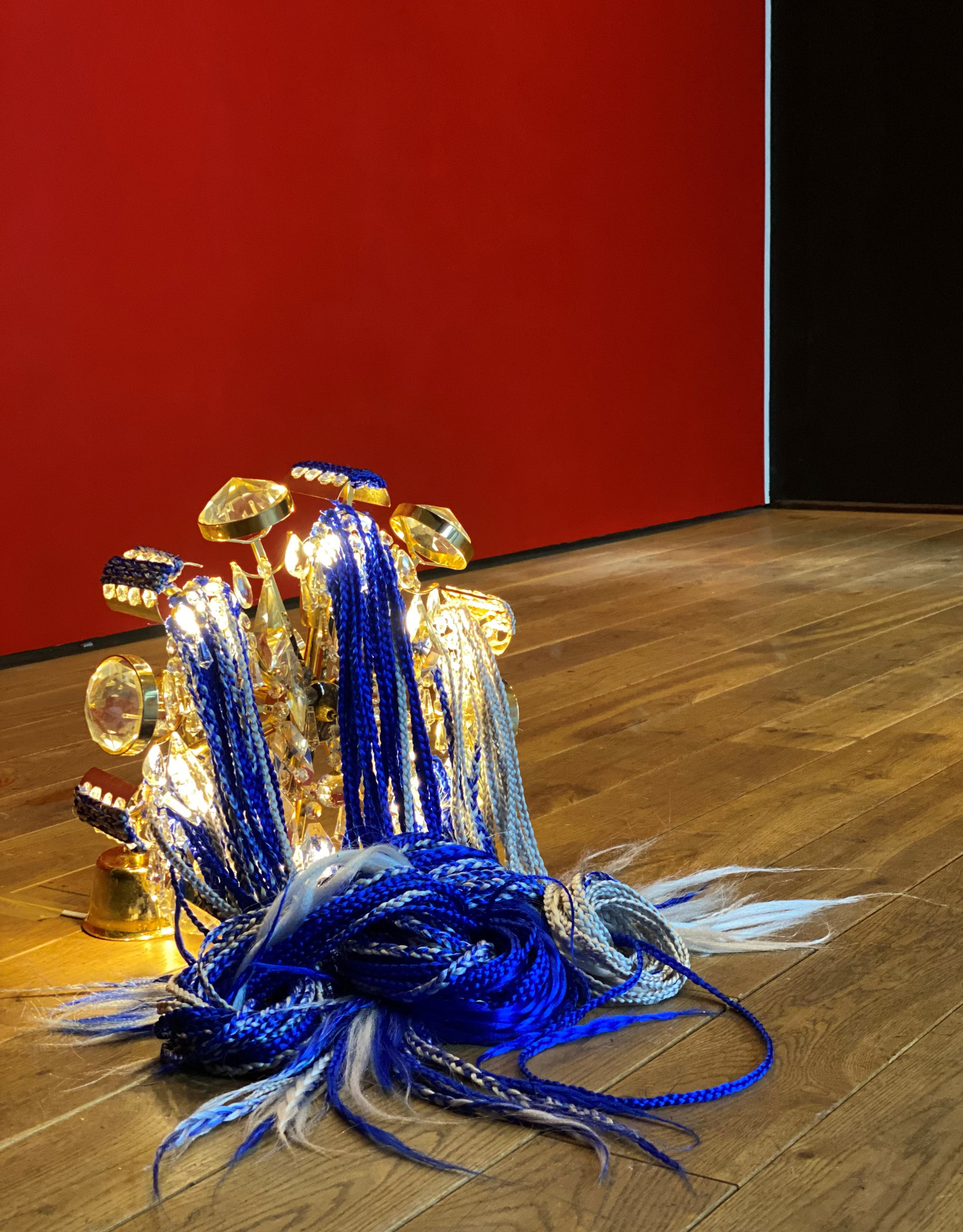 Image of an artwork placed on a wooden floor with a red wall behind it. The sculpture consists of glowing golden lamp and partially braided synthetic hair in diffrenet tones of blue