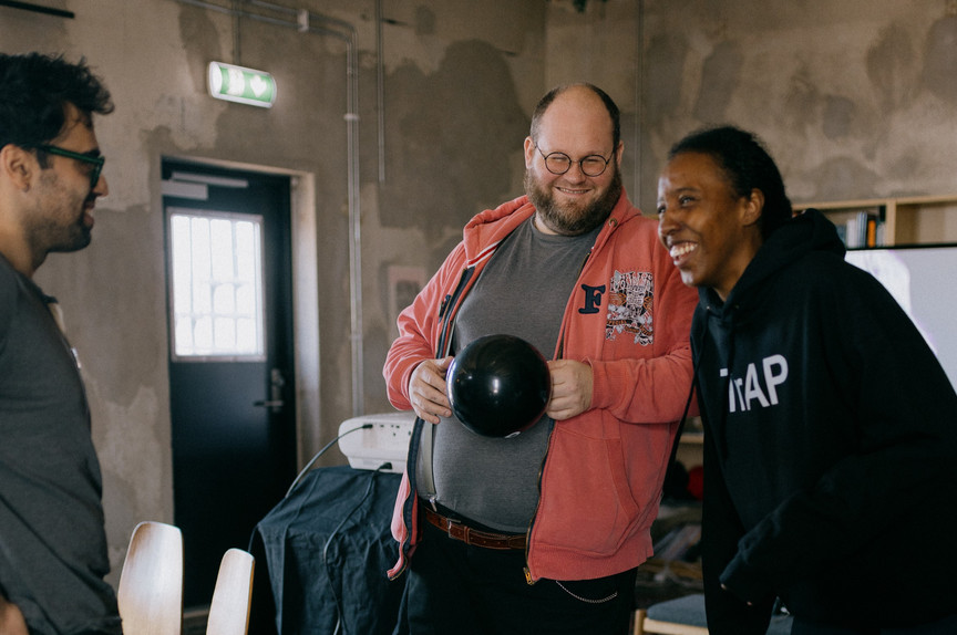 Three people are smiling and looking at a shiny, black sphere held by the person in the middle. The man in the center is wearing glasses, a gray t-shirt, and a reddish jacket with a logo on the left side of the chest. He also has a brown belt. The woman on the right is wearing a black hoodie with white letters on the left side of the chest. The man on the left has dark curly hair and glasses, and he is wearing a dark gray t-shirt. They are standing in a room with gray walls, a door with a window on the left, and some equipment covered by a dark cloth under the middle man. There are also chairs in the room. The atmosphere appears light and friendly.
