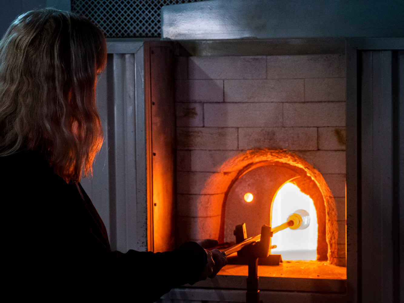 We see the back of a longhaired individual holding a metal rod with liquid glass at the end. There's a bright orange glow from the kiln.