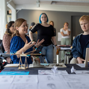 A medium shot shows a group of six people gathered around a table covered with papers and models. The focus is on a woman with shoulder-length brown hair and glasses, wearing a blue patterned top, who is gesturing with both hands as she speaks and smiles. She is seated behind the table, slightly to the left of the center. To her right, a young man with red hair is also seated at the table, looking towards the woman with a neutral expression. He is wearing a dark blue zip-up hoodie over a white t-shirt and has a tablet resting on the table in front of him. A blue paper cup with a white lid sits near his hands.

Behind the woman in the blue top, another woman with blue hair and a black shirt stands, looking towards the right side of the frame. Further back, two more women are partially visible, standing and looking towards the table. To the left of the woman in the blue top, a woman with dark hair and a patterned shirt is visible from the shoulders up, looking towards the speaker. A black backpack rests on a chair to the left of the frame.

The table is covered with a dark cloth and holds various papers with drawings and diagrams, as well as small architectural models made of light-colored materials. The background shows a brightly lit room with white walls, pillars, and a glimpse of a window on the far left. The overall atmosphere suggests a workshop or presentation setting.