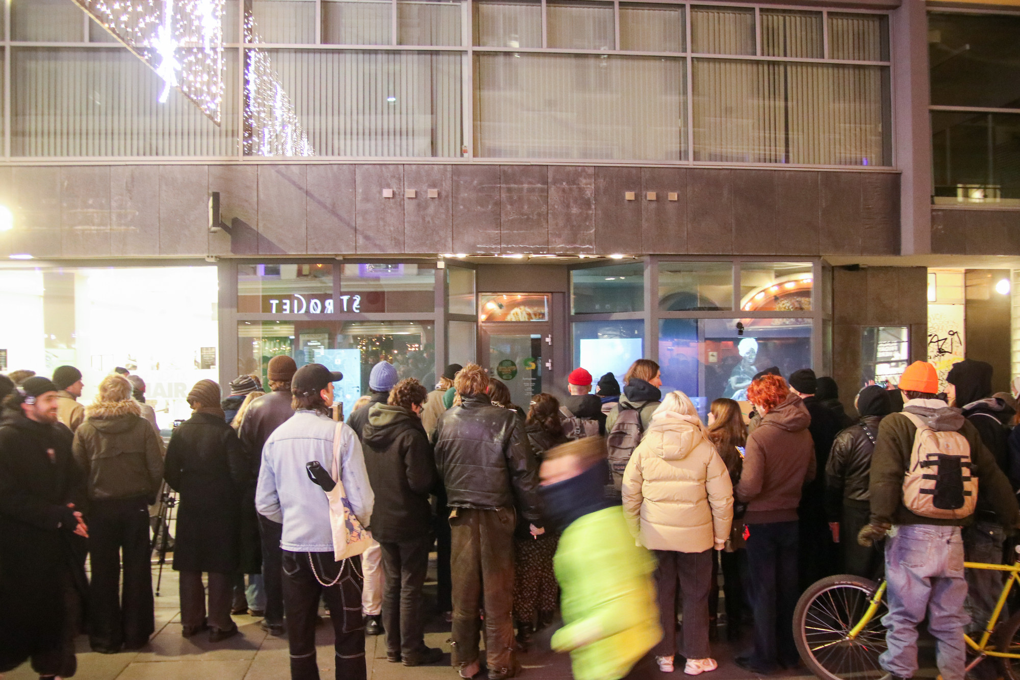 Around thirty people are standing with their backs towards the photographer on the sidewalk in front of a building. They are looking through the display windows at videos projected on the walls inside the premises.