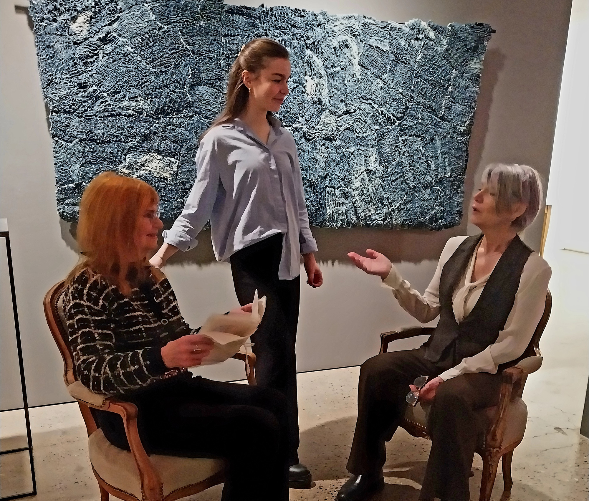 Photo of three people. Two women sitting in armchairs half facing eachother. Between them a woman standing. The grayhaired woman to the right is talking and gesticualting with one hand. The redhaired woman on the left is holding a paper. Her and the standing woman are looking at the person talking.
Behind them on the wall there is a gray blue textile art piece hanging. 