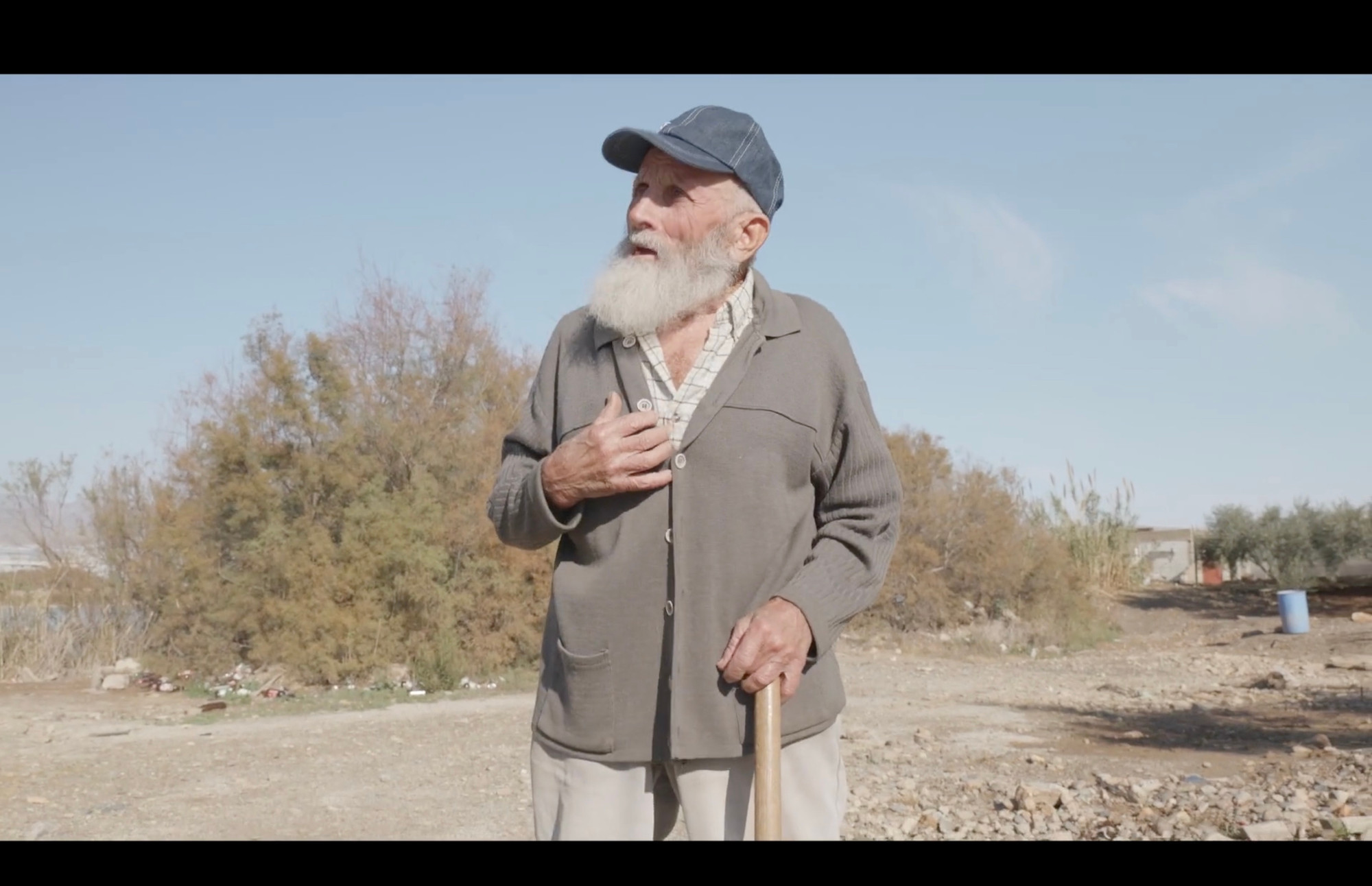 Still image from film– an old man with a cap and a beard standing in the middle of the image. In the background some dry land with bushes and light blue sky