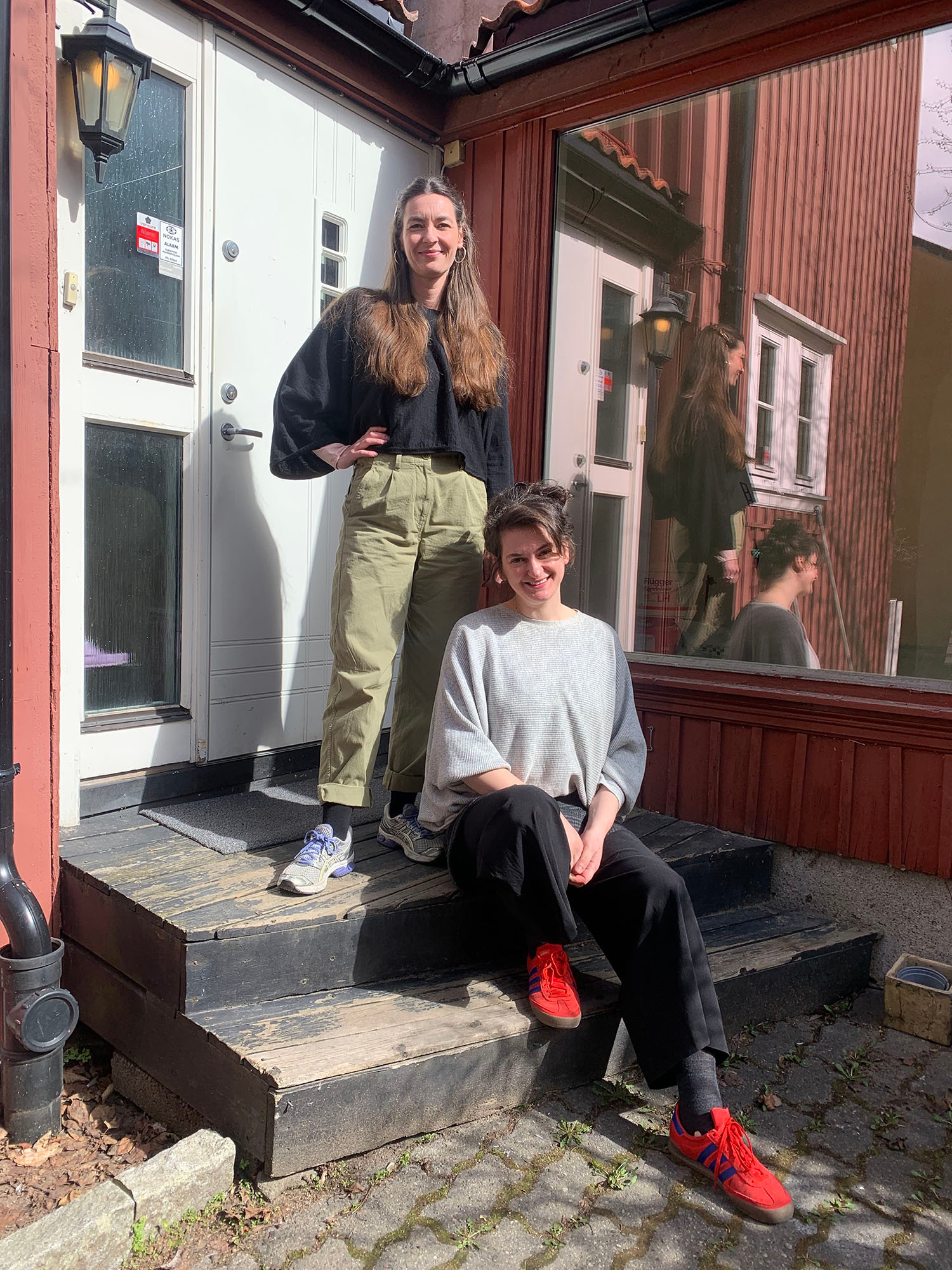 Two women on the a doorstep of a red wooden building. Sophie is standing on the top step, and Jasmina is sitting in front of her.