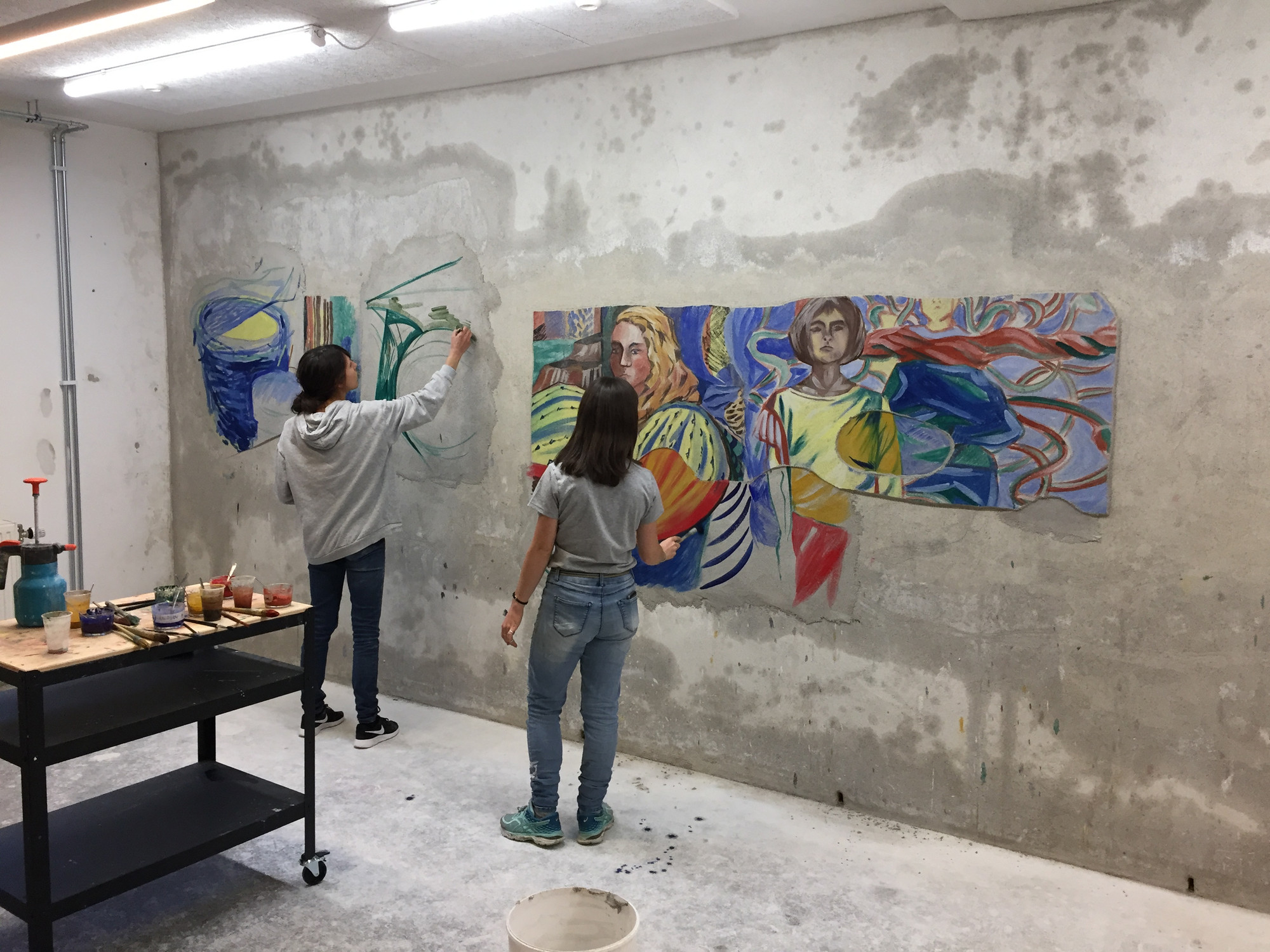 A gray room where two people– Mari Kolbeinson and Laura Gaiger– are making paintings directly on one of the walls. Behind them there is a trolly with cups of paint and painting tools.