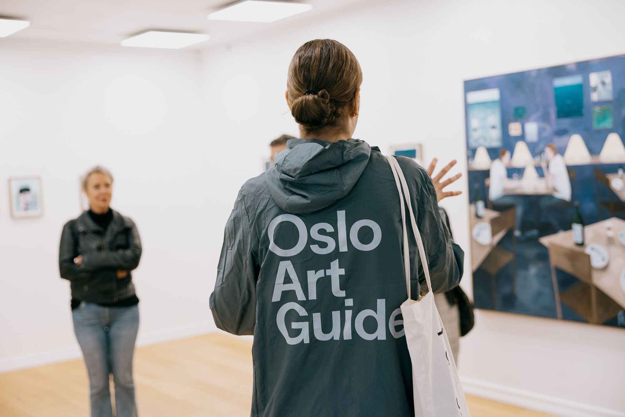 En kvinne har på en jakke med teksten Oslo Art Guide. Hun snakker til to tilhørere foran et maleri.