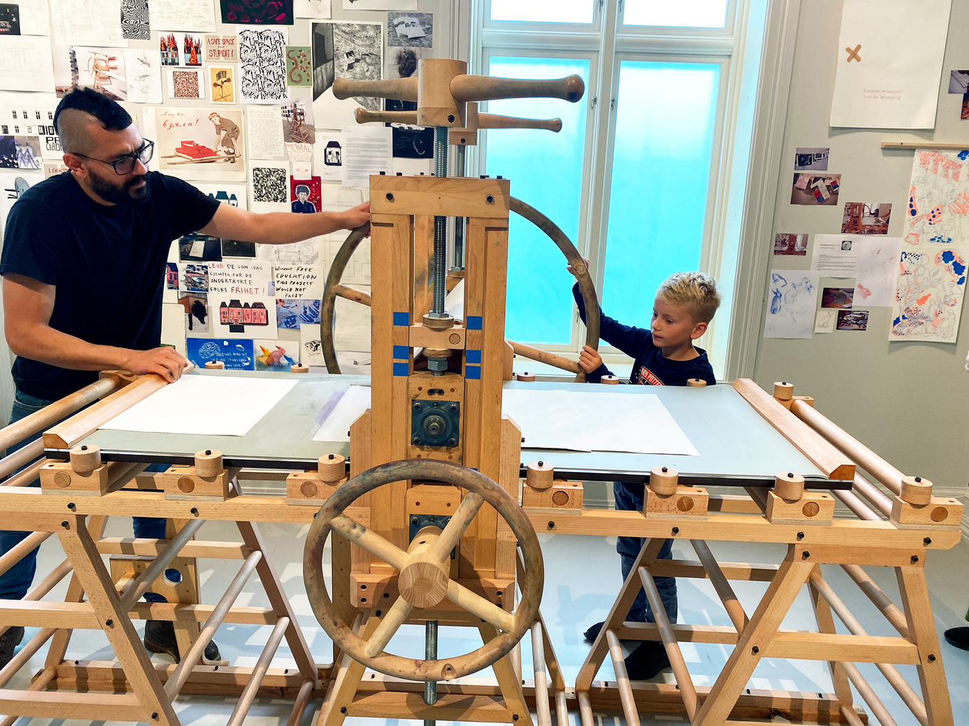 An adult and a child turn the wheel on a large wooden printing press