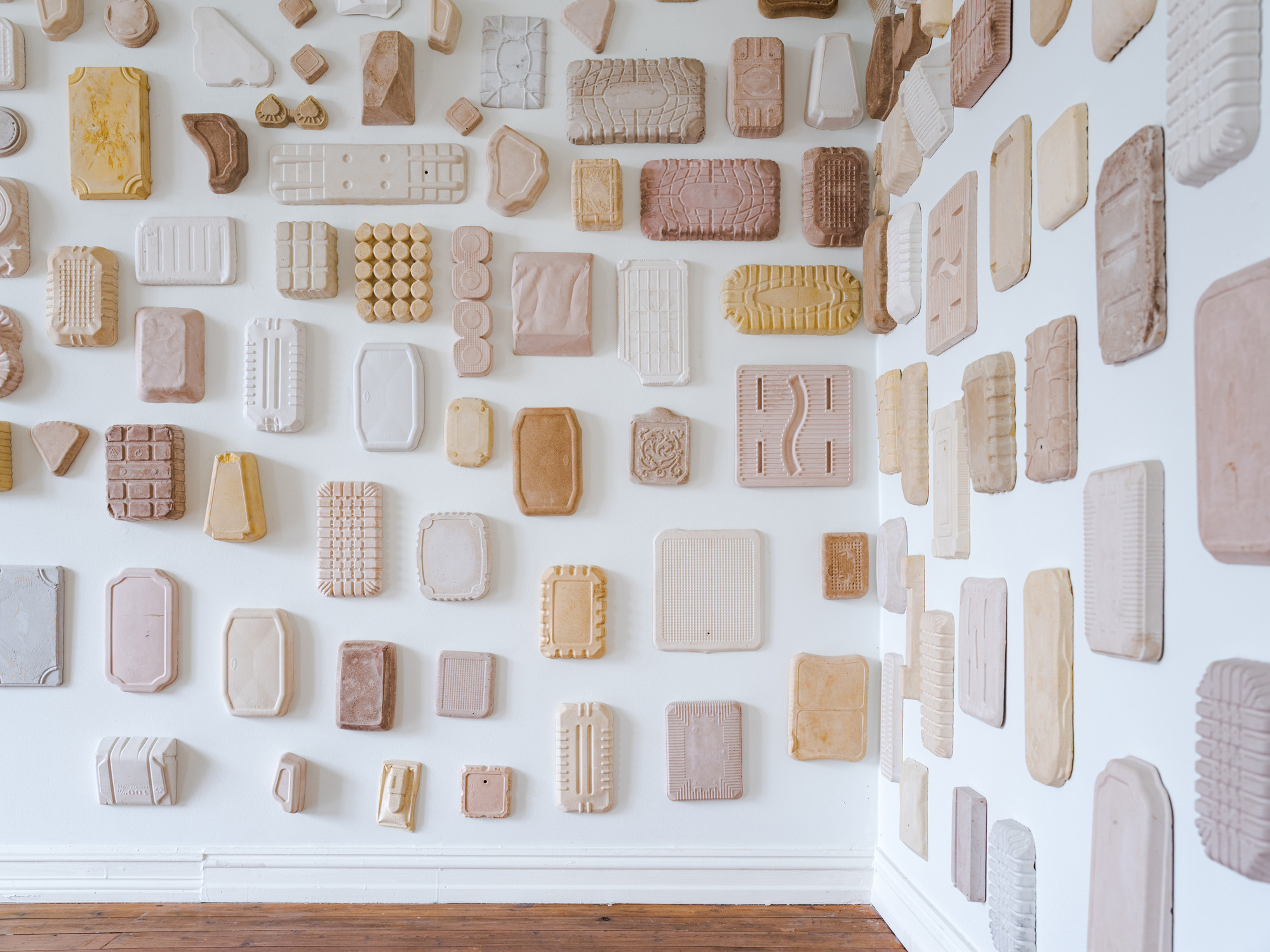 The corner of a room where the two visible walls have around 80 abstract plaster sculptures in different shades of white, yellow and red attached to them. 