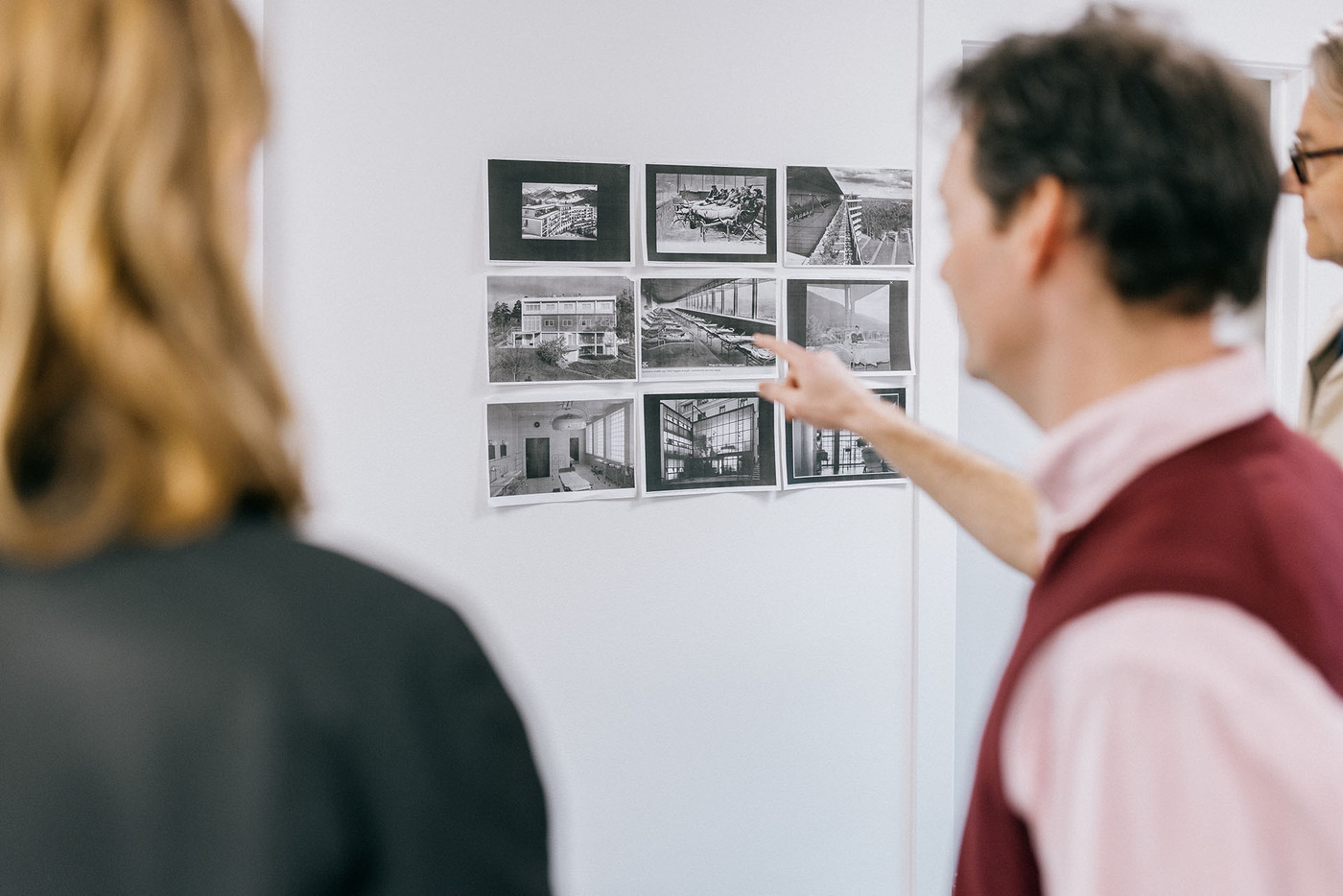 A man with short, messy hair, is standing between two visitors, and pointing to black and white photographies on the wall.