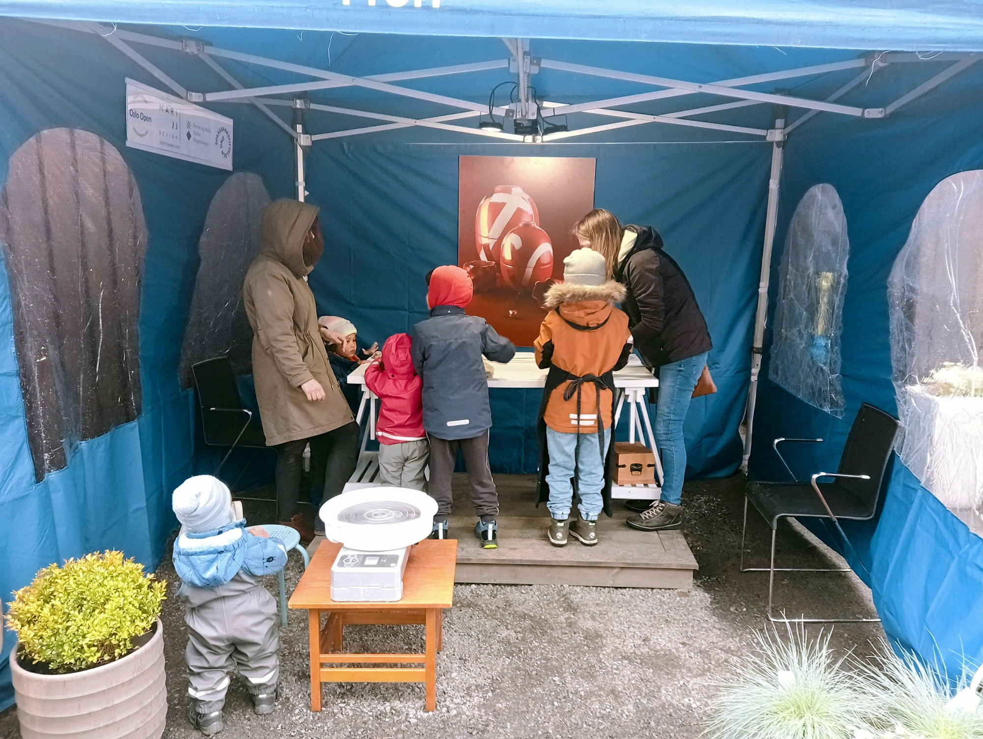 The inside of a blue party tent that is open on one side. Furthest in there is a table with two adults and four kids around it. Closest to the camera there is a large flower pot, a toddler and a small table with a pottery wheel.