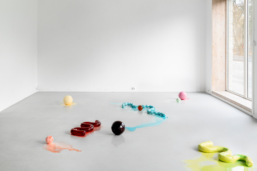 Nine abstract sculptures in different colors and shapes lie on the floor of a gallery space. The colors of the sculptures are: warm yellow, pink, mint green, amber, turquoise, red, black, peach and yellow-green. The sculptures are either spherical or have wavy shapes. Small puddles flow out of some of the sculptures. The gallery space has white walls, a light gray floor and a large window facing the street.