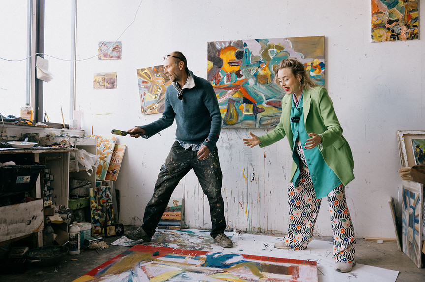 Artist Jens Hamran and reporter Ragnhild Brochmann leans over a canvas on the floor. Paint stains, clutter and paintings in rich colours fills the space.