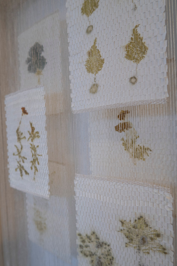 Detalje-bilde av verket Herbarium. Her vises nærbilde av hvit silke med plantetrykk som er klippet opp i strimler og vevd i firkanter i A5 format. Silken har klare trykk av blomster og blader i duse toner av gult, grønt og blått.