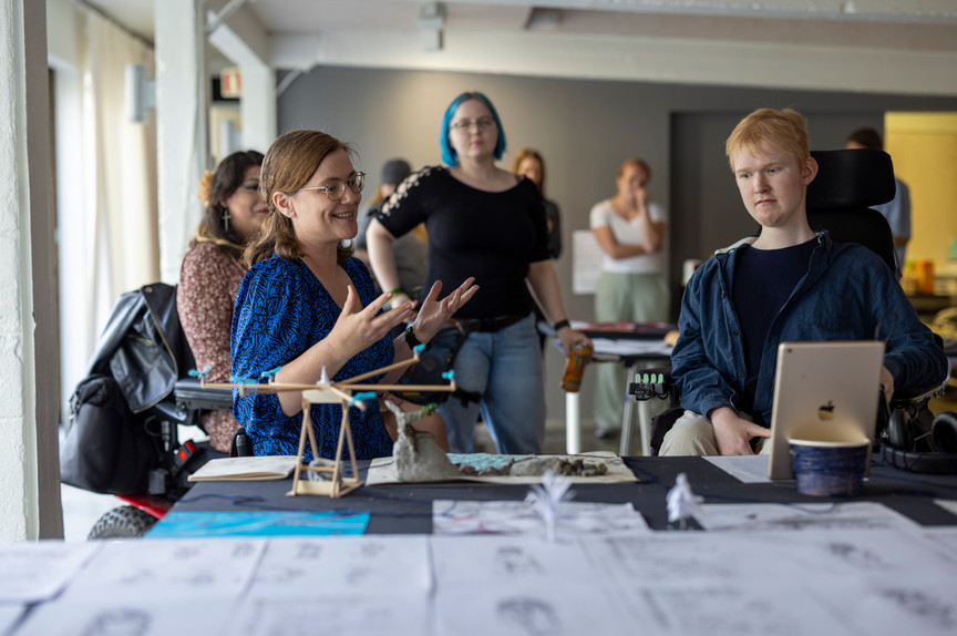 A group of people are gathered around a table covered with papers and models. The focus is on two people in front, a young woman and a young man, who are both seated in wheelchairs. The woman is gesturing with both hands as she speaks and smiles. 