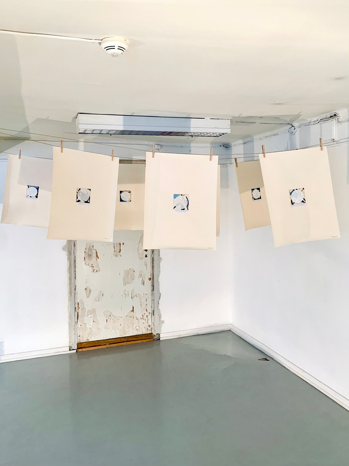 Flere store løse ark i natur-toner med mindre collage-motiv svever i et gallerirom, hengende på klessnor med klesklyper