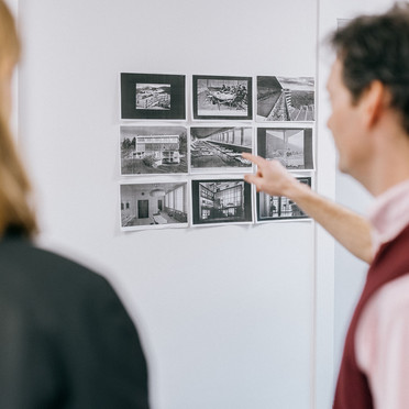 A man with short, messy hair, is standing between two visitors, and pointing to black and white photographies on the wall. 