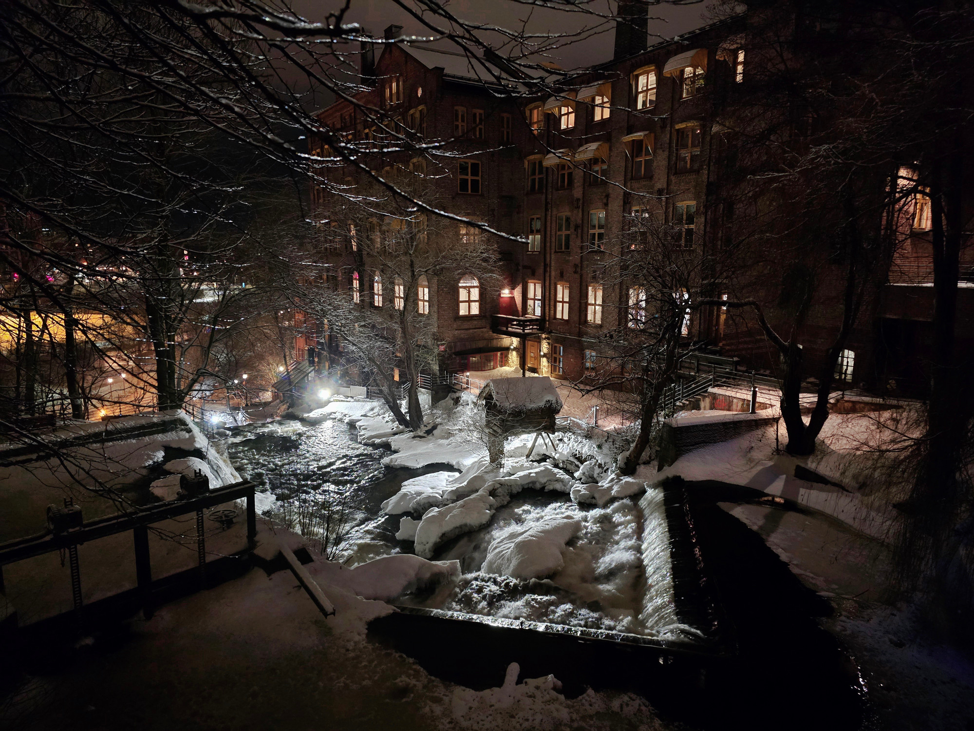 A dark city view during winter with snow on the ground. There is a river running through with some trees and both banks. In the background a large brick building with five floors. Some of the windows are glowing with light.