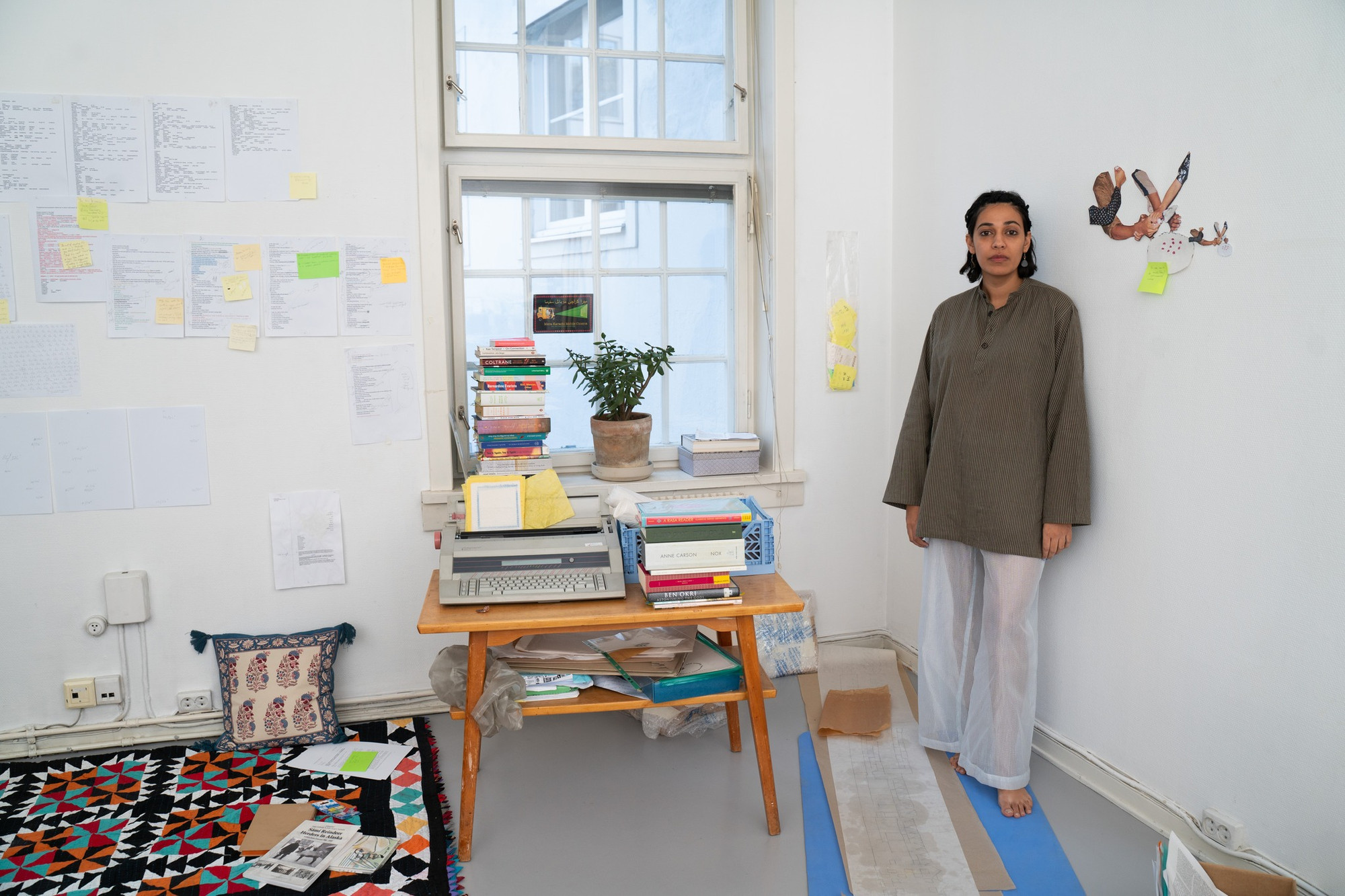 The artist standing in a tidy studio with papers and post its covering the walls.