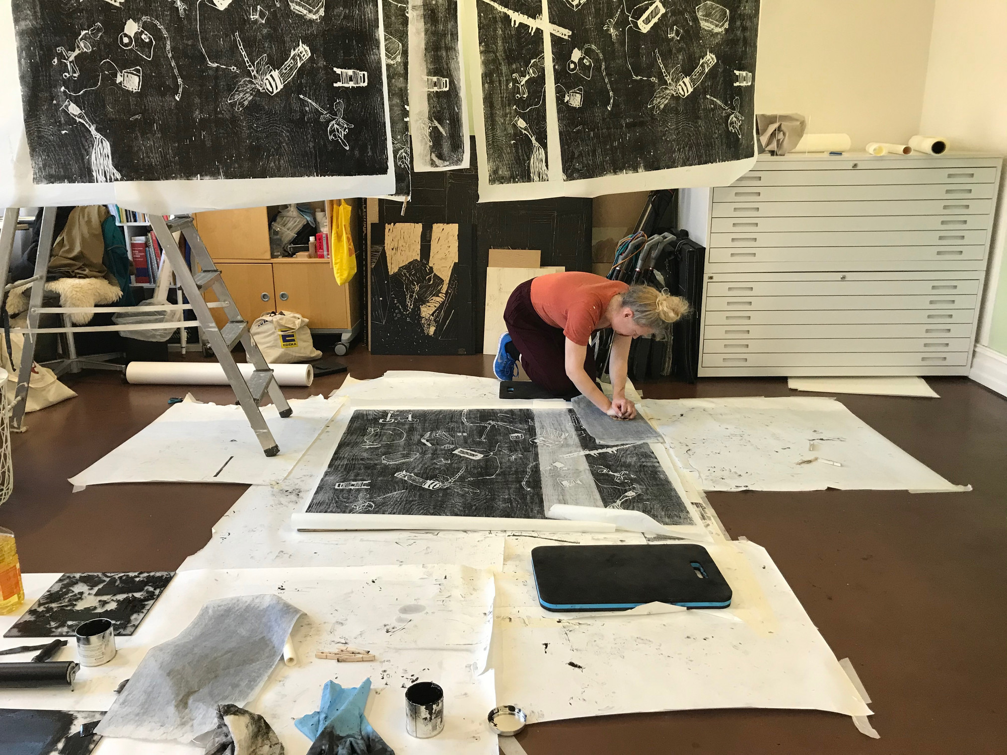 Photo from an artist studio. The artist is working kneeling on the floor. There are large black and white prints hanging from the ceiling and placed on the floor. In the foreground there are toold and materials for printing.